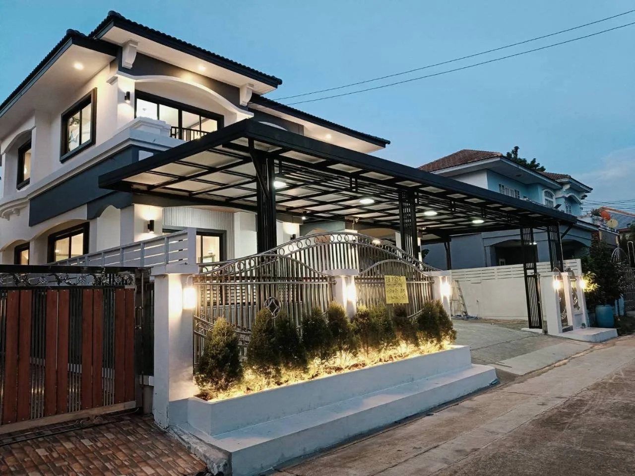 ด้านหน้าบ้านเดี่ยว 2 ชั้นตอนพลบค่ำ มีโรงจอดรถหลังคาโครงเหล็กและแปลงไม้ประดับริมรั้ว